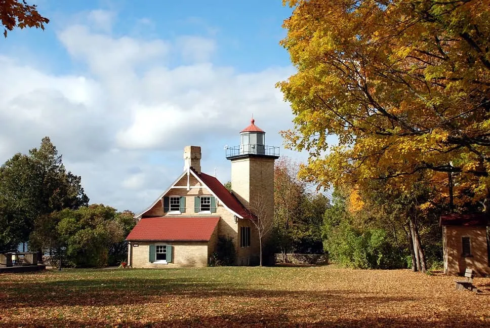 Door County attractions in September, View of Eagle Bluff Lighthouse with single tower and small living quarters nearby with trees in fall colors to either side and a lawn of green grass partially covered in fallen leaves in front
