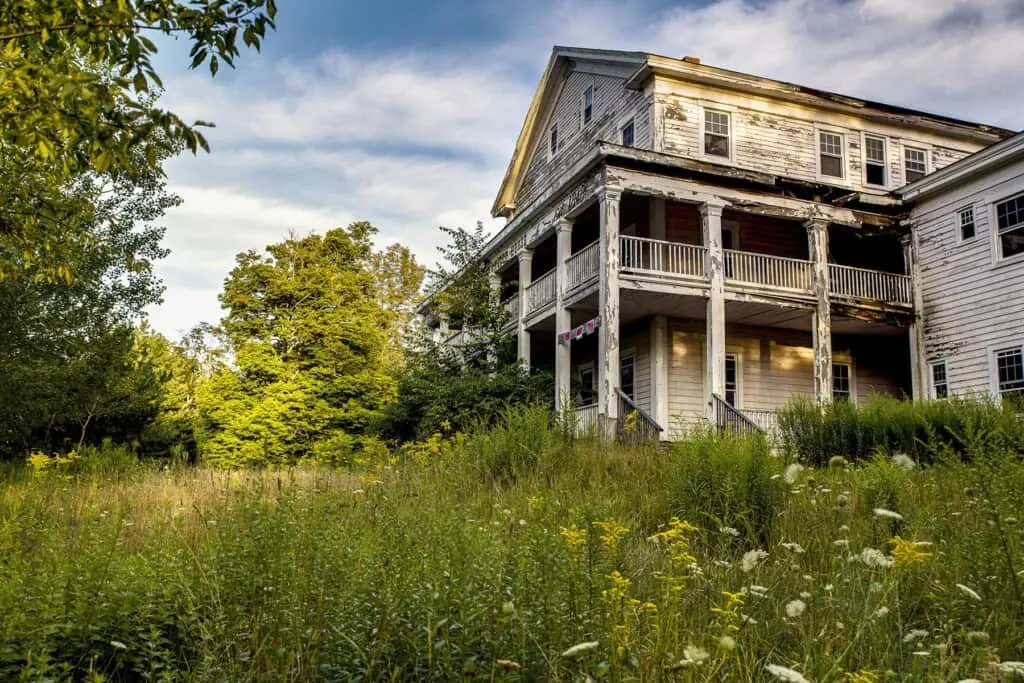 Explore these deserted places in usa, large white painted wooden house with wide covered porch and overgrown green garden