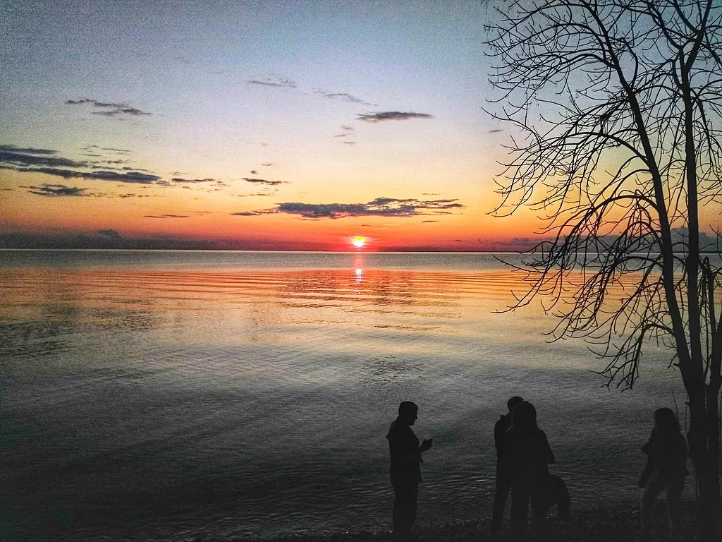 people hanging out lakeside at low sunset with the bright red sun reflected in the still waters of the lake in the distance