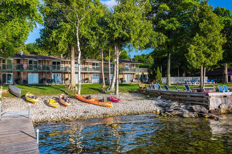 the shallows resort with wooden jetty next to stony beach with several kayaks resting next to green trees and grass with a series of apartments behind