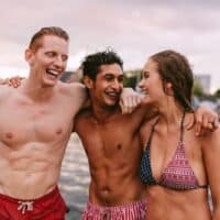 Shot of young friends in swimwear standing together by the lake and laughing. Men and woman enjoying a day by the lake.