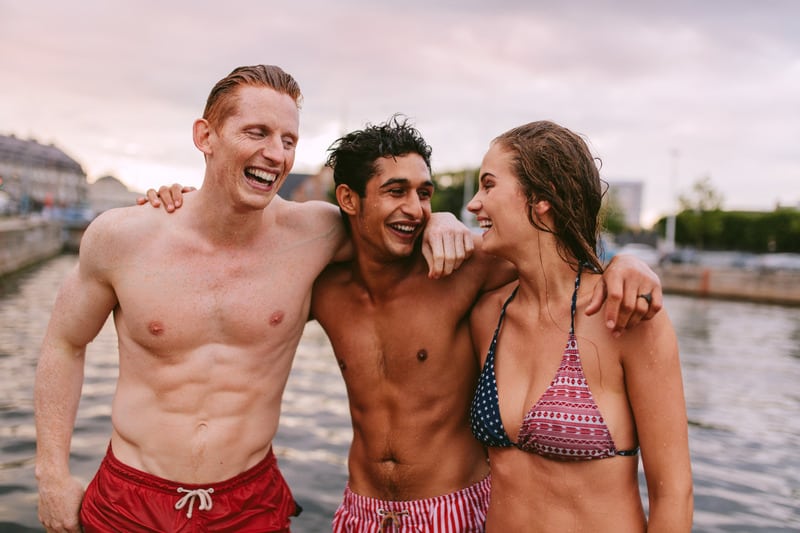 Shot of young friends in swimwear standing together by the lake and laughing. Men and woman enjoying a day by the lake.