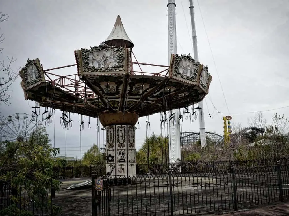 Find the most abandoned places in america, tall ruined fairground ride with broken detailing and faded paint scheme with large abandoned rollercoaster behind
