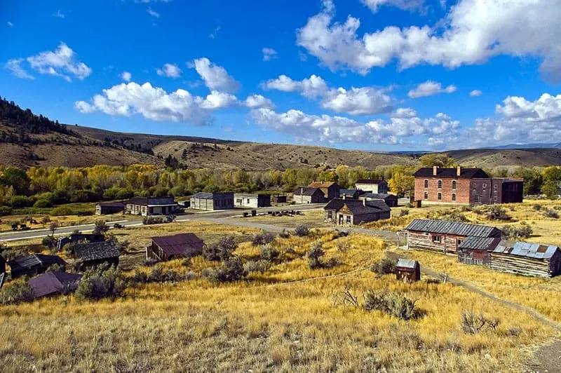 Travel to the most deserted place in USA, aerial view of ghost town with several abandoned wooden houses and cabins in a green valley with rolling hills behind under a blue sky with fluffy clouds