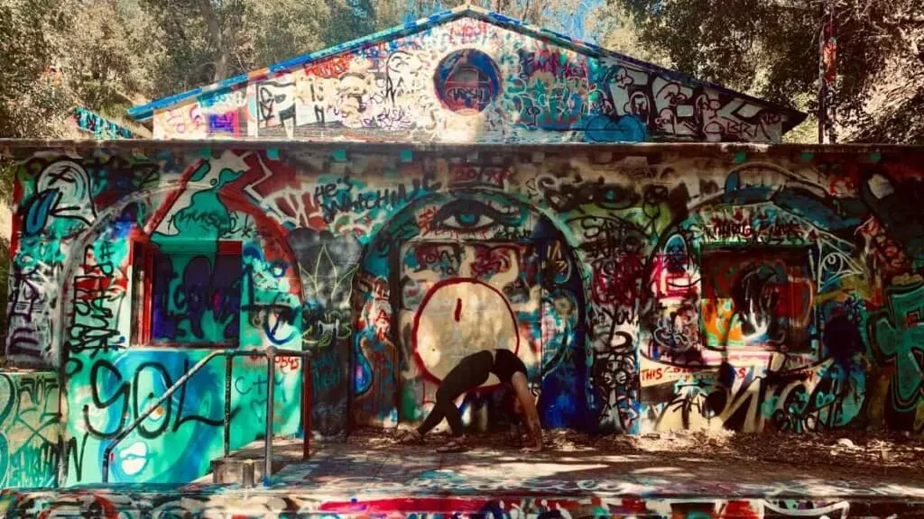 Wander among the abandoned places in the united states, person holding yoga position in entranceway of large abandoned house covered in colourful graffiti under a canopy of trees in the sunshine