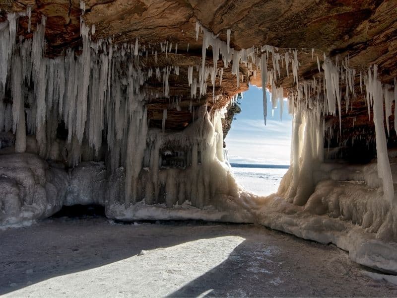 best winter places in Wisconsin, Apostle Islands Ice Caves on frozen Lake Superior, Wisconsin with frozen shards of ice hanging down from the cave ceiling and a hole in the wall looking out onto the wide open lake covered in snow