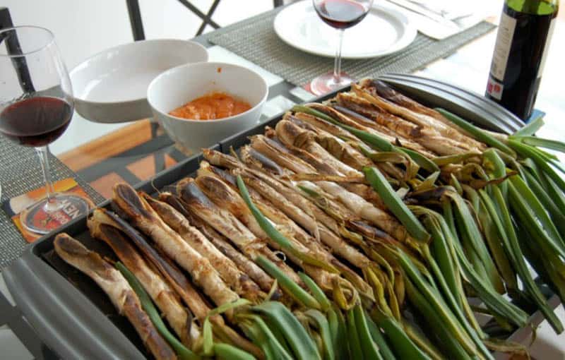 Calçots dish with a row of cooked Calçots sitting on a metal tray with plates and glasses of wine nearby