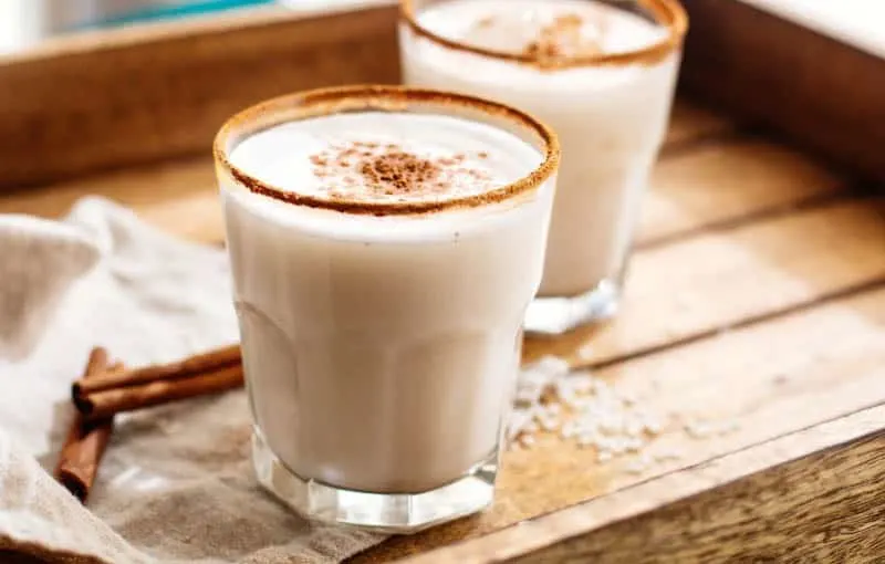 Horchata Drink in small tumbler glasses sitting on a wooden tray with some cinnamon sticks nearby