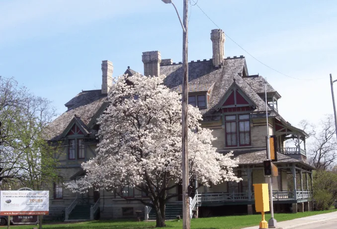 Top Places to Visit in Appleton, Wisconsin, Front view of Hearthstone Historic House Museum with white tree on lawn in front and welcome sign to one side under a bright blue sky