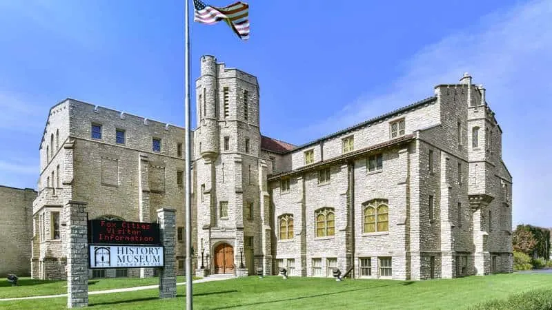 Best things to do in appleton, Wisconsin, History Museum At The Castle building made of grey stones with large central turret and two wide wings with vaulted windows and flagpole with the USA flag in the foreground all under a bright blue sky