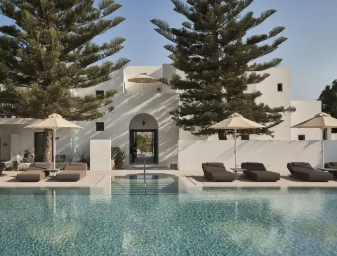 pool with front view of hotel with pool side beds, umbrellas, and pin trees with a building and a door in the middle