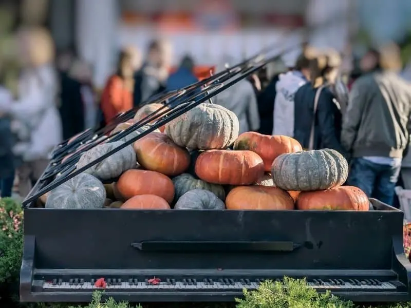 Spend October in Wisconsin this year, old piano with lid open on top of a pile of pumpkins in orange and grey colors with crowds of people visible in the distance behind