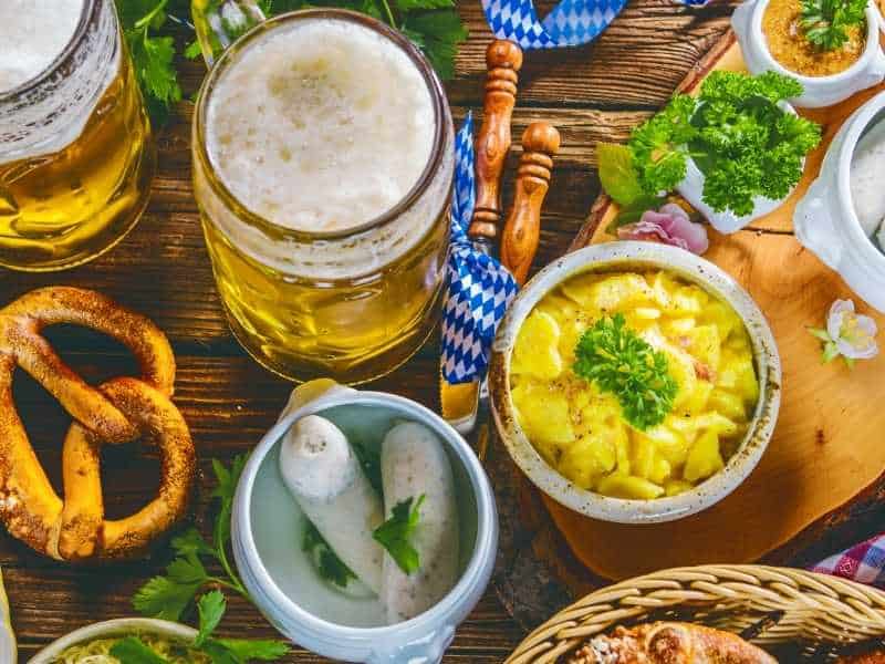 These top Wisconsin breweries are well worth a visit, shot of a wooden table covered in various foodstuffs including a pretzel, a ceramic bowl with two sausages, some green herbs, some chopped potatoes and a pair of large glasses of amber beer