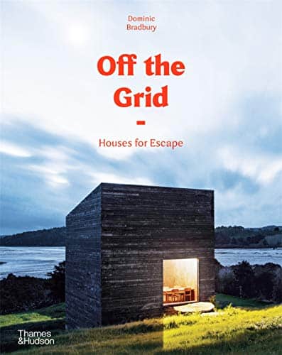 Coffee table book travel, book cover showing very modern wooden and glass cabin at dusk with view inside showing wooden table and chairs with a backdrop of a lake and some hills in the distance