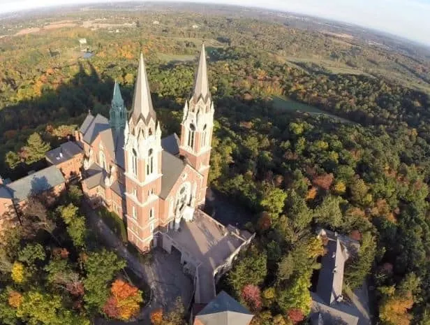 Cool things to do in milwaukee in october, aerial view of holy hill church in milwaukee with its two tall pointed towers surrounded by a forest of trees in fall colors