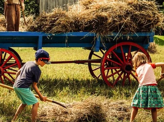 Enjoy the delights of October in Milwaukee, two small children picking up hay with pitchforks with a wagon full of hay visible behind them