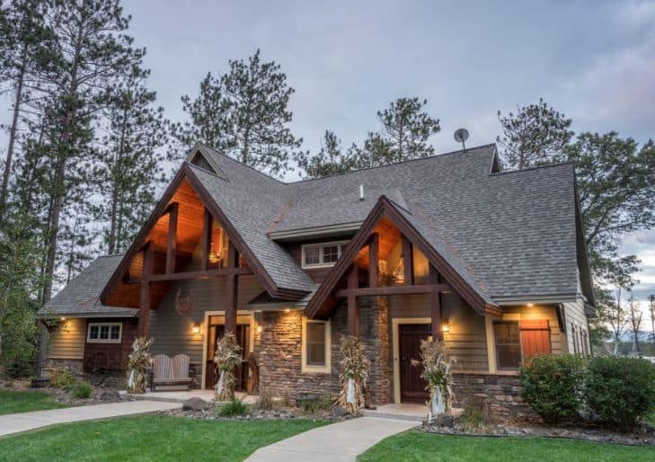 Best Luxury Cabins in Northern Wisconsin for fall stays, front view of Spectacular Luxury Lakefront Cabin with large sloping rooftops in grey tile underlit by warm orange lights with decorative plant arrangements attached to the supporting wooden pillars with a row of thin tall trees behind on a grey cloudy day
