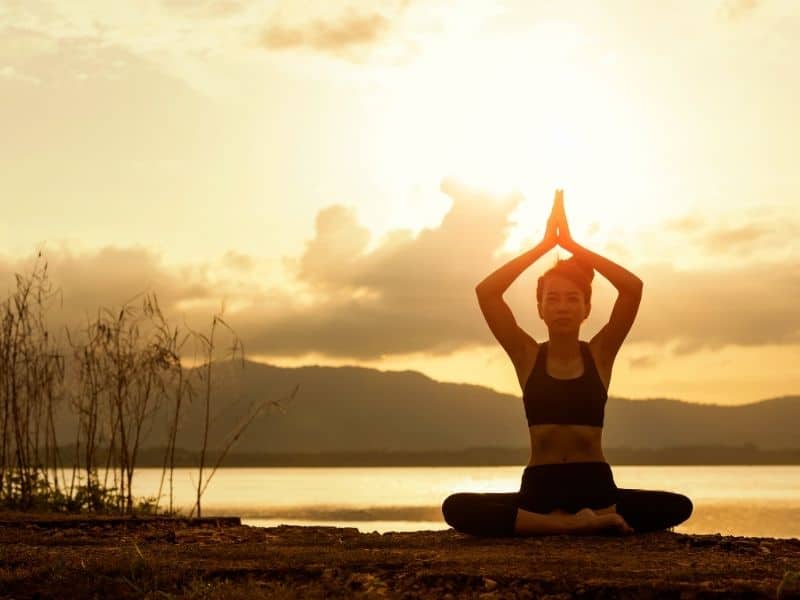 best brands for sustainable yoga clothes, woman meditating in yoga clothes next to a lake at sunset