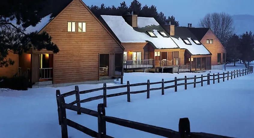 a wooden lodge with roofs and terrace covered in snow, snow field with gate and trees at the back