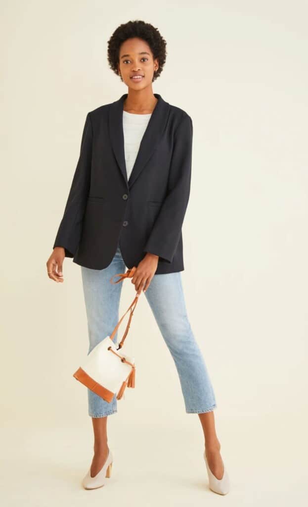 brands that use recycled materials, black woman wearing white shirt under black suit top with mid-calf length jeans and white heels, she is carrying a white bag