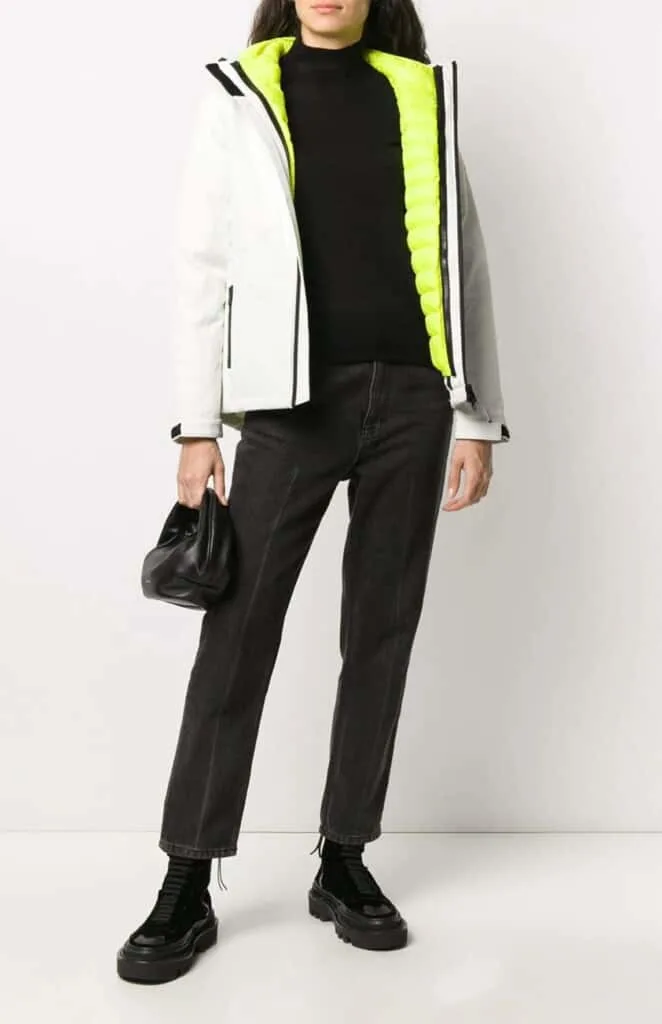 recycled material clothing brands, image of androgynous person from chin down wearing long sleeve white coat with neon lining, black t-shirt, black jeans and black boots, they are holding a black purse
