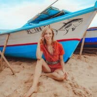 eco fashion blogger wearing recycled red romper sitting in the sand next to a blue and white boat with a fish painted on the side