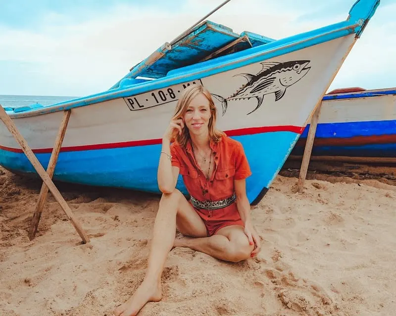 why buy from recycled clothing brands, eco fashion blogger wearing recycled red romper sitting in the sand next to a blue and white boat with a fish painted on the side