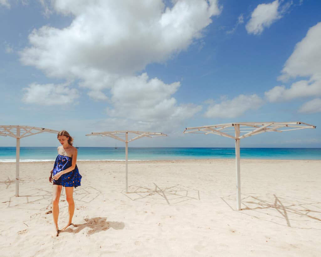 best recycled clothing brands, fashion eco blogger walking in a blue romper through a sandy beach next to empty umbrella holders under a cloudy blue sky with clear blue water in the background
