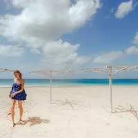 best recycled clothing brands, fashion eco blogger walking in a blue romper through a sandy beach next to empty umbrella holders under a cloudy blue sky with clear blue water in the background