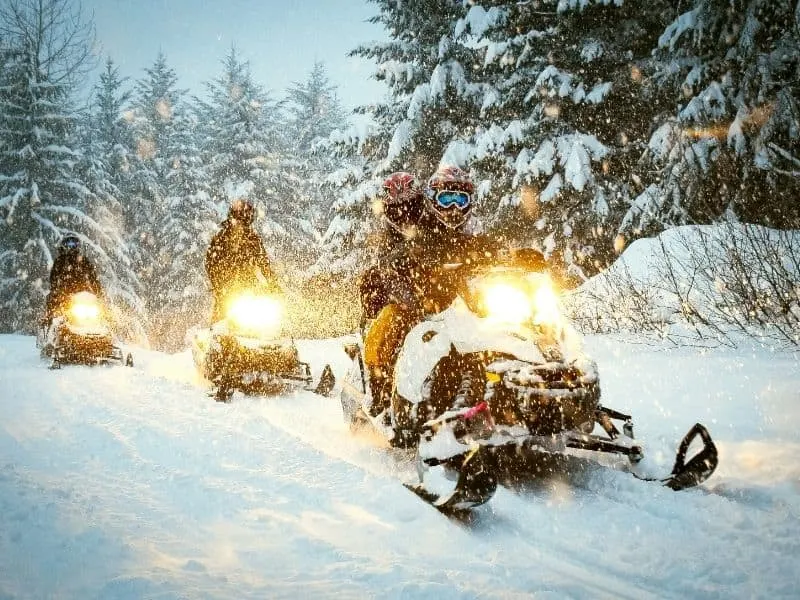winter activities in Wisconsin Dells to enjoy in January, three snowmobiles riding through the snow at the snow at low light with bright yellow lights on each