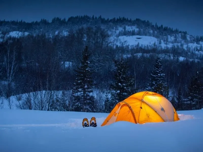 Best Sites for Winter Camping in Wisconsin, yellow tent in the snow at night light up from the inside with snowy mountain and trees in background