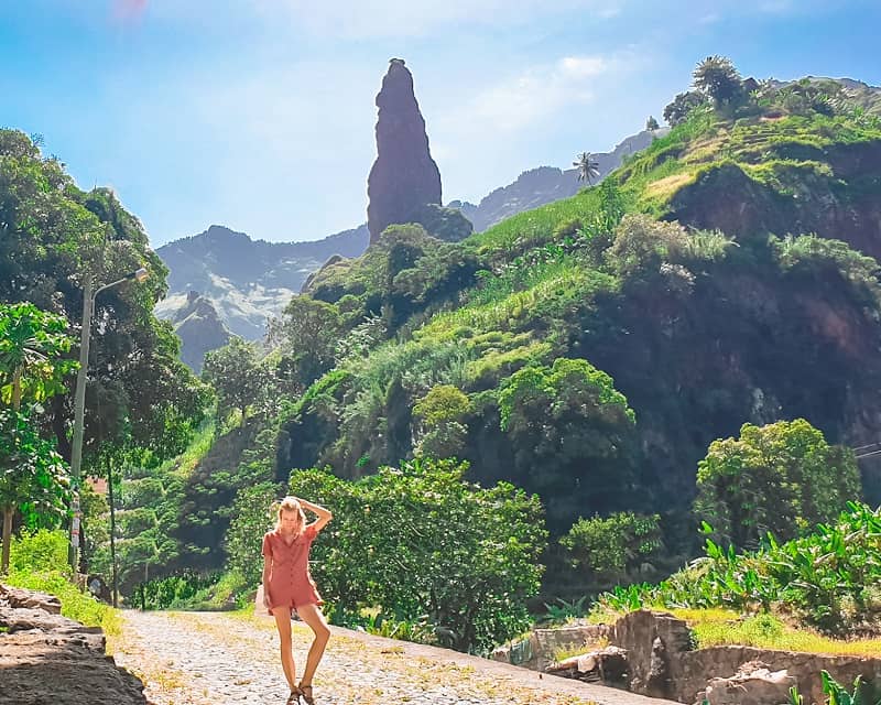 a woman on a pathway filled wtih mountains and lush vegetation with a protruding rock at the back