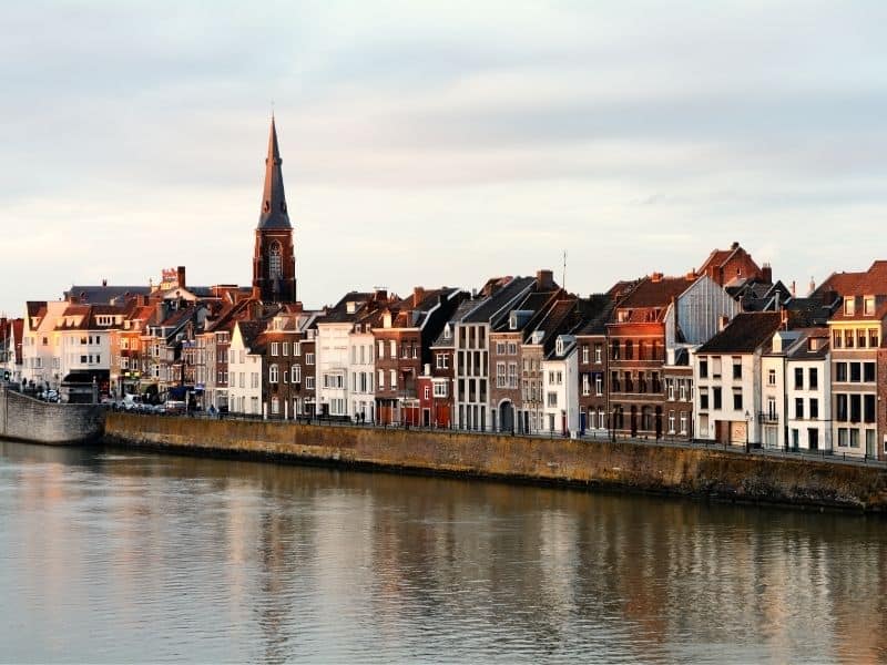 Head to these places to visit close to Luxembourg, view from the side of a river of the densely packed residential townhouses of maastricht netherlands with a tall church tower to one side all under a cloudy sky