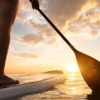 Stand up paddle boarding on quiet sea, legs close-up, sunset