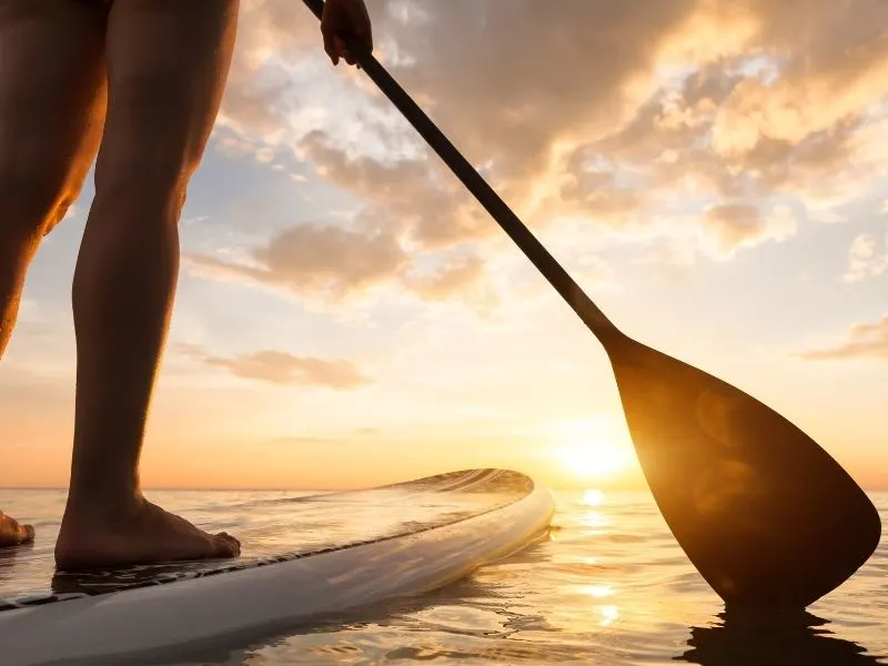 Stand up paddle boarding on quiet sea, legs close-up, sunset