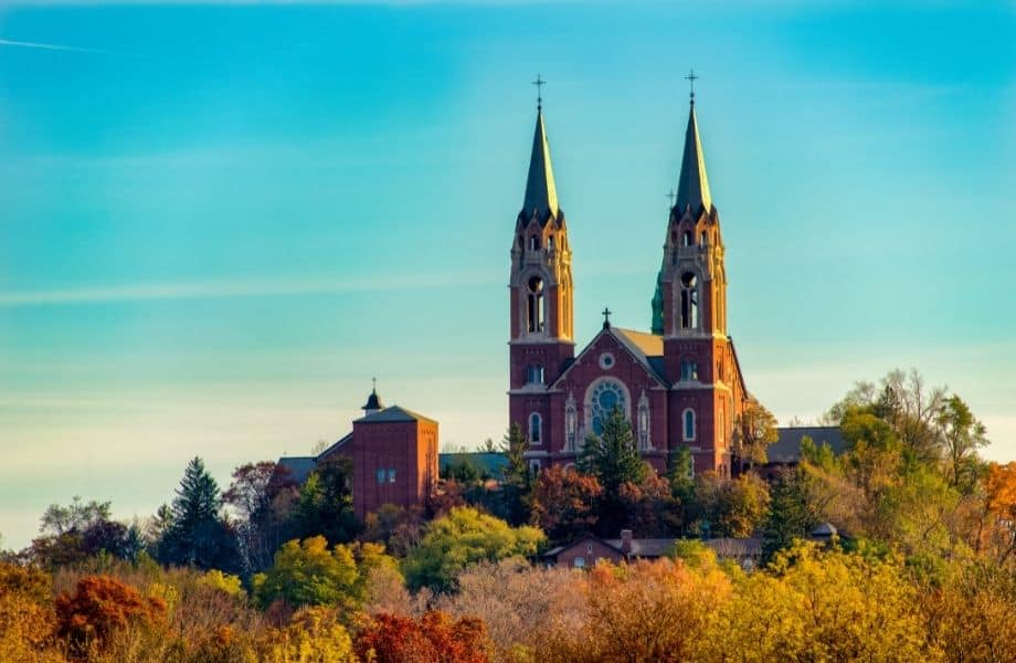 view of the Holy Hill National Shrine of Mary with its two large pointed towers sitting atop a hill covered in trees bearing fall colors all under a bright clear sky