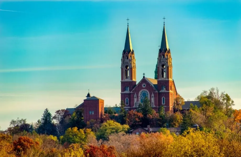 Find the best places to see fall colors in wisconsin, view of the Holy Hill National Shrine of Mary with its two large pointed towers sitting atop a hill covered in trees bearing fall colors all under a bright clear sky