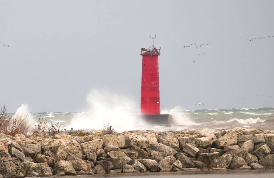 Sheboygan Lighthouse a red lighthouse being hit by waves with rocks on the forefront on a dark day with birds flying. where to stay in Sheboygan