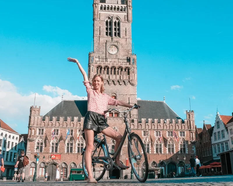 rent a bike in bruges, belgium, a woman on a bike in front of a building with a clock tower in bruges