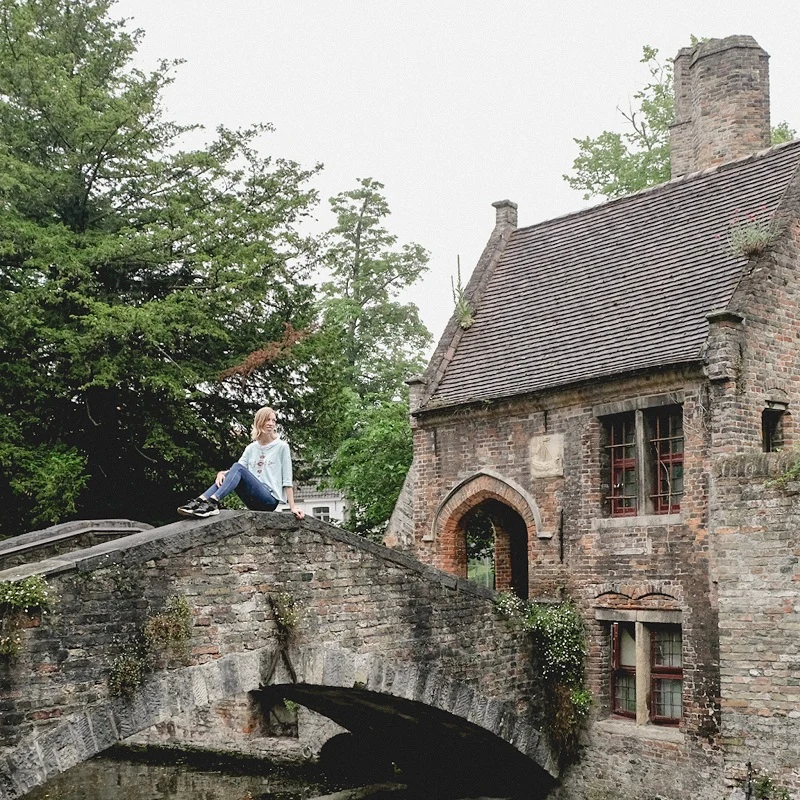 a woman sitting on lovers bridge bruges belgium with a stone house on the background