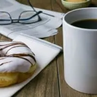 best cafes in Milwaukee, coffee mug next to a donut on a napkin and folded up newspaper with glasses on top of a table