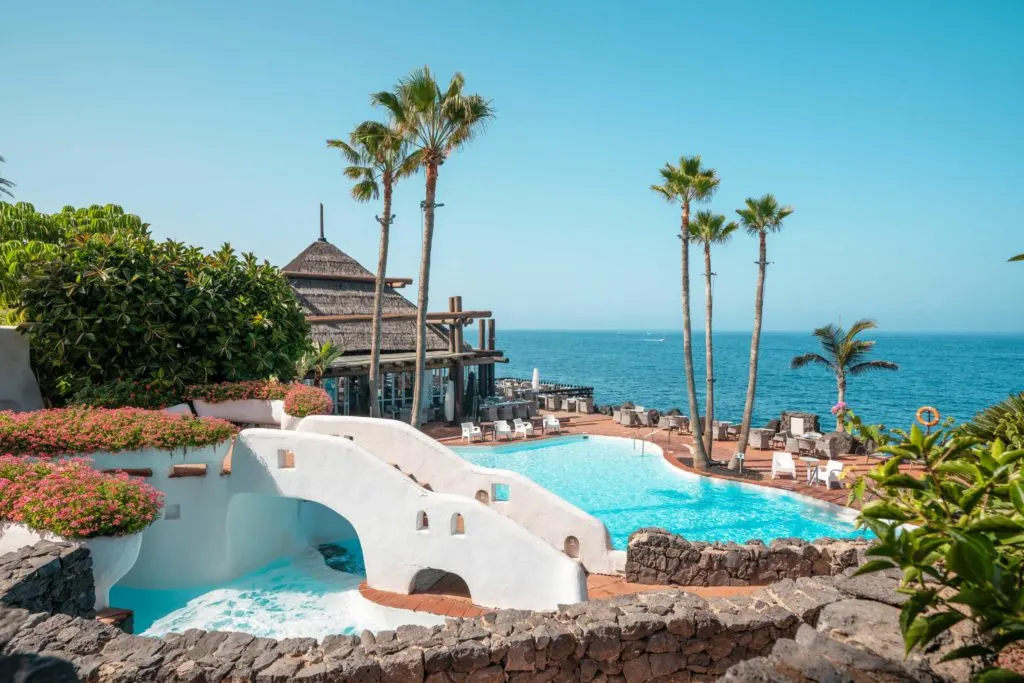 Visit the best beachfront hotels in Costa Adeje, outside pool and seating area with palm trees overlooking the ocean