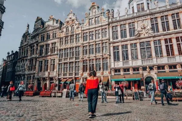 a woman in red standing center with the grand palace of brussels in the background as well as a crown