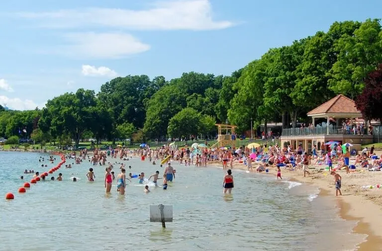 many people on Riviera Beach in Lake Geneva with trees at the back under a blue sky with clouds