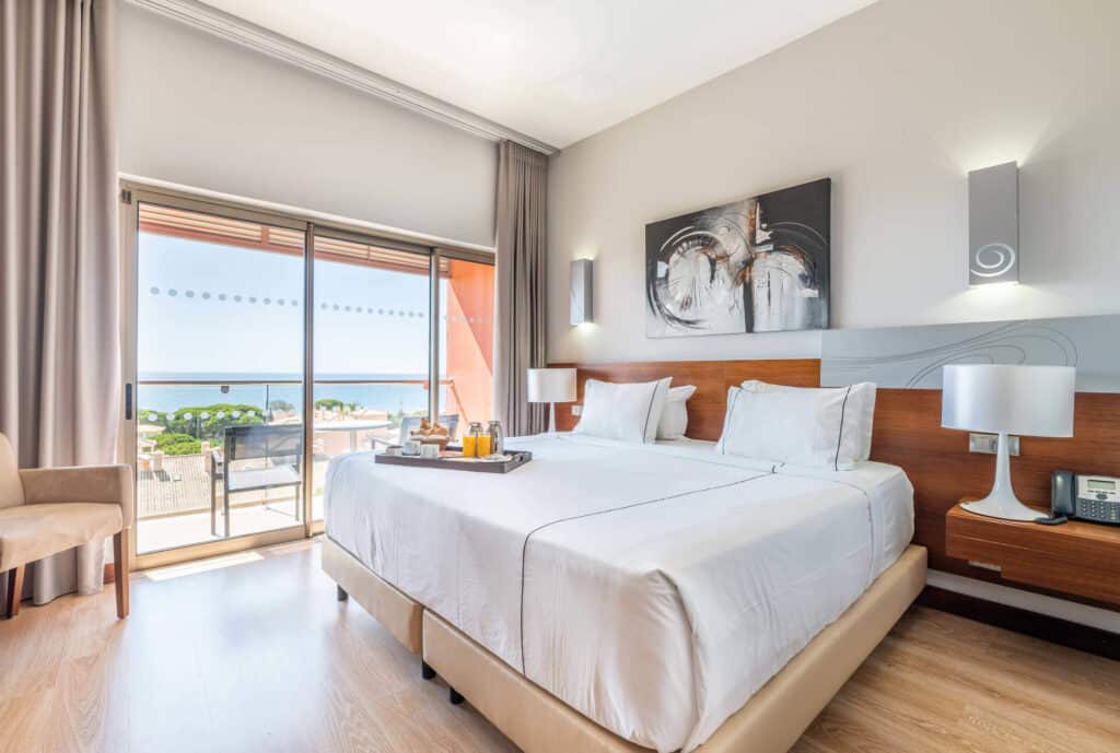 best places to stay in Albufeira, hotel room with large bed with a food tray on top, a painting on the wall and a balcony with a view of the beach on a sunny day