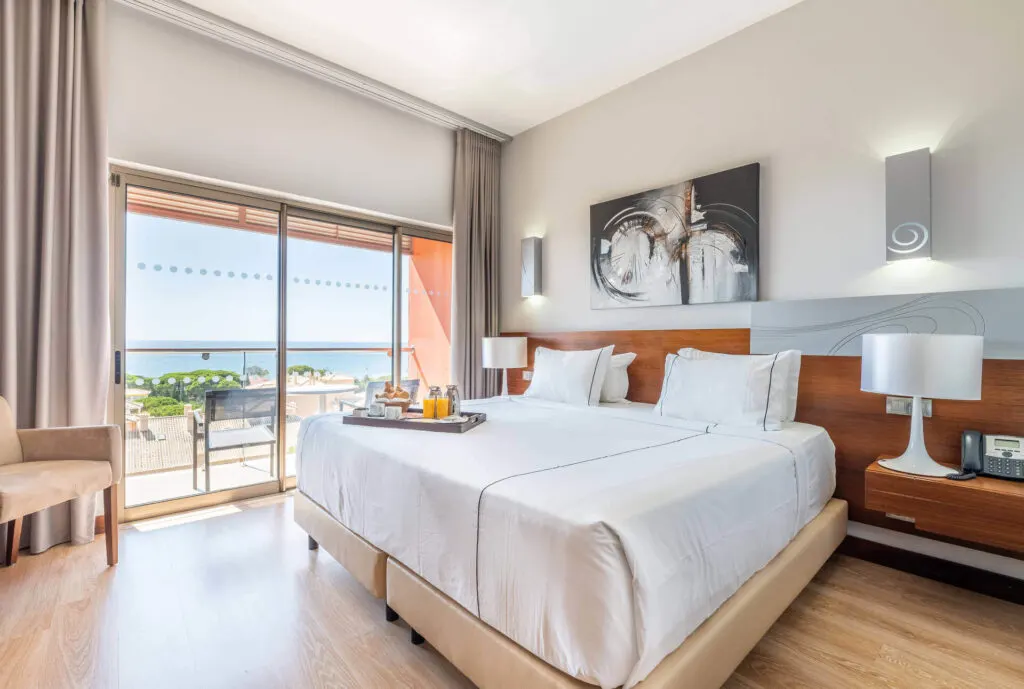 best places to stay in Albufeira, hotel room with large bed with a food tray on top, a painting on the wall and a balcony with a view of the beach on a sunny day