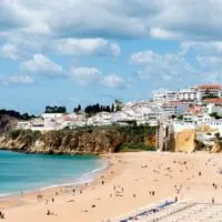 Best Hotels in Albufeira Algarve, sandy beach sparsely populated with holidaymakers next to blue waters with white foam surrounded by short cliffs with cluster of white-walled buildings overlooking the bay under a cloudy blue sky