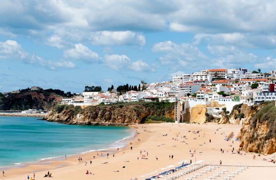 Best Hotels in Albufeira Algarve, sandy beach sparsely populated with holidaymakers next to blue waters with white foam surrounded by short cliffs with cluster of white-walled buildings overlooking the bay under a cloudy blue sky