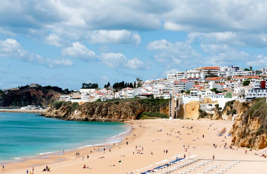 View of Fisherman's Beach in Albufeira, sparsely populated with holidaymakers next to blue waters with white foam surrounded by short cliffs with cluster of white-walled buildings overlooking the bay under a cloudy blue sky