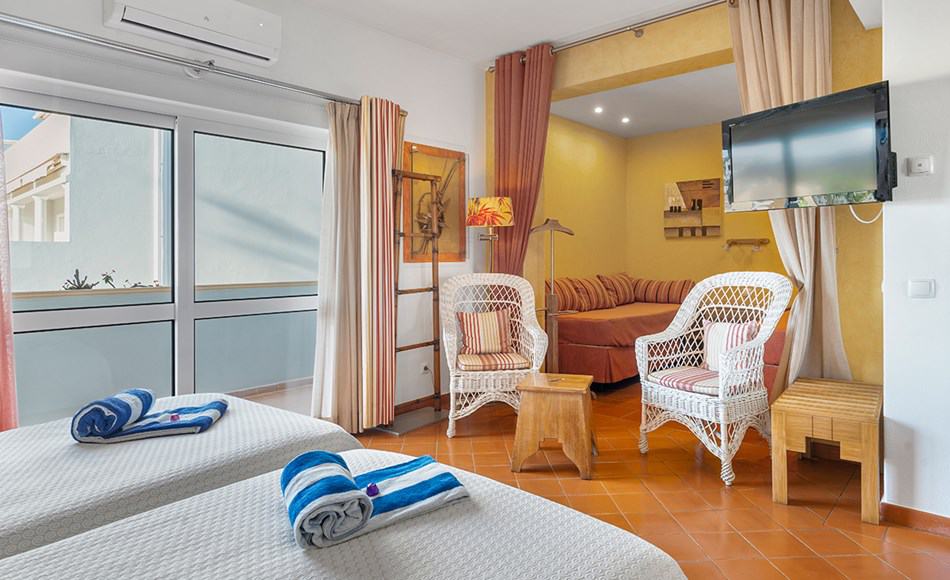 best hotels in Albufeira, hotel room with two beds with rolled towels and chocolates on top, a TV and two wicker chairs and an alcove with a wide couch/day bed